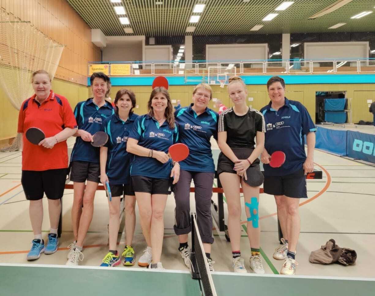 Gruppe von sieben Tischtennisspielerinnen in Sportkleidung, stehend vor einem Tischtennistisch in einer Halle.