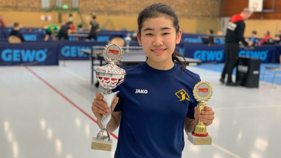 Junge Sportlerin hält zwei Trophäen in der Hand, im Hintergrund eine Tischtennis-Spielhalle mit Spielern.