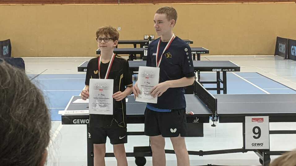 Zwei Tischtennisspieler stehen mit Urkunden und Medaillen auf einem Tischtennisturnier, Tischtennisplatten im Hintergrund.