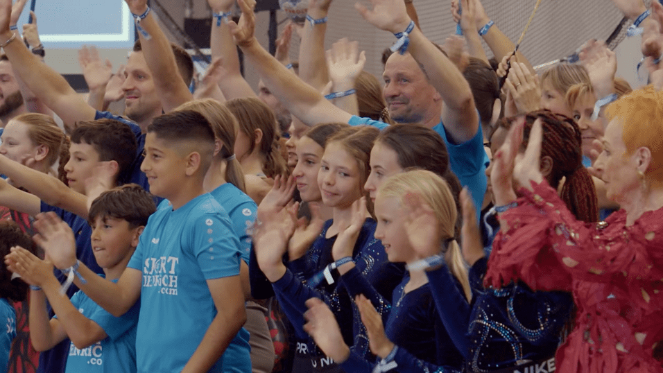 Gruppe von Kindern und Erwachsenen in blauen T-Shirts, die begeistert applaudieren und lächeln.