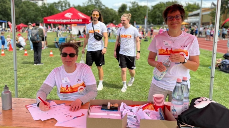Zwei Personen an einem Tisch mit Materialien und Wasserflaschen, während im Hintergrund Teilnehmer eines Sportevents zu sehen sind.