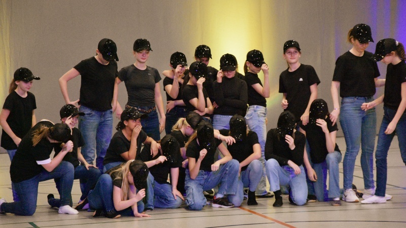 Tanzgruppe in Schwarz und Jeans mit glitzernden Caps, posierend in einer Sporthalle.