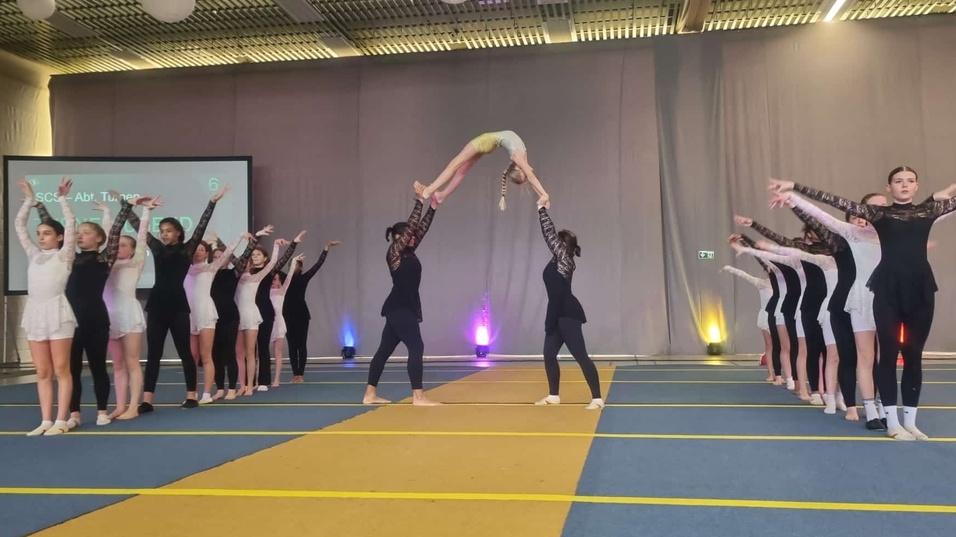 Tänzerinnen in Sportkleidung führen eine Hebefigur auf einer Bühne während einer Aufführung aus.