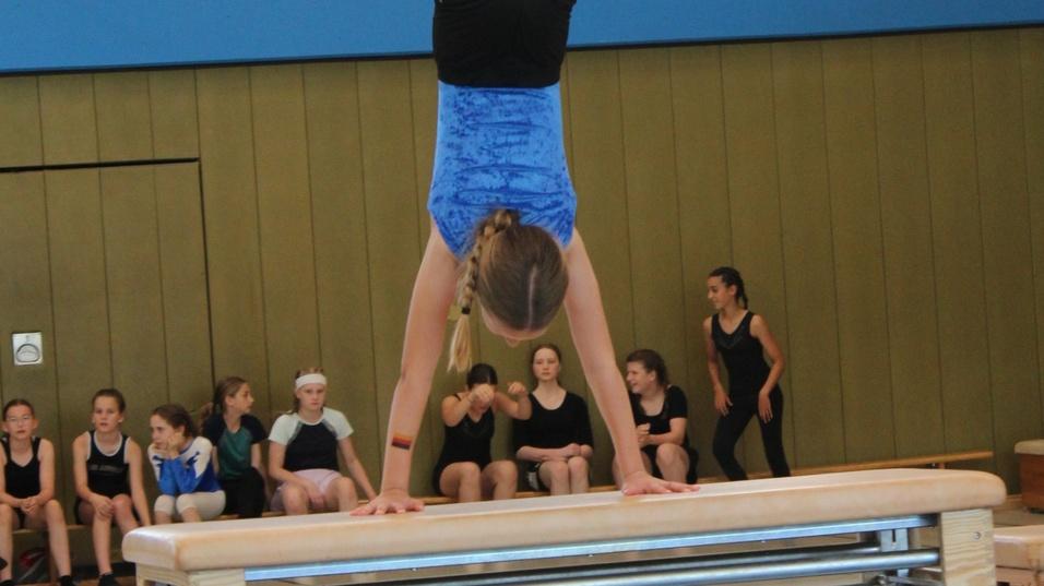 Mädchen übt eine Handstandübung auf einem Barren, während andere Jugendliche zuschauen.