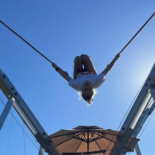 M&auml;dchen schwingt kopf&uuml;ber an einem Seil unter blauem Himmel &uuml;ber einem Sonnenschirm.