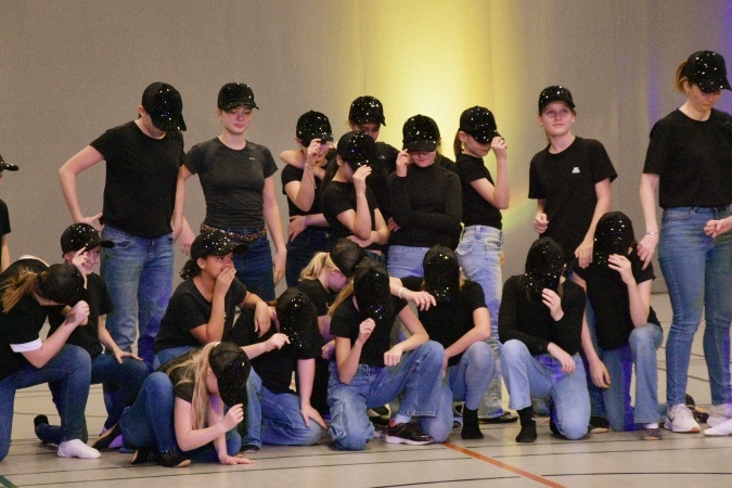 Tanzgruppe in Schwarz und Jeans mit glitzernden Caps, posierend in einer Sporthalle.