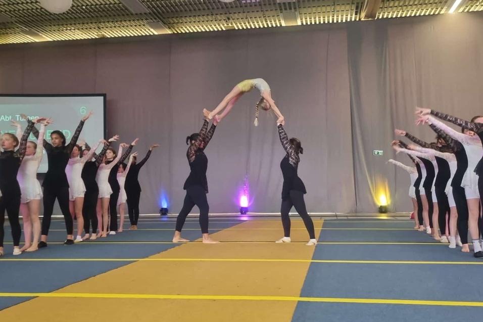 T&auml;nzerinnen in Sportkleidung f&uuml;hren eine Hebefigur auf einer B&uuml;hne w&auml;hrend einer Auff&uuml;hrung aus.