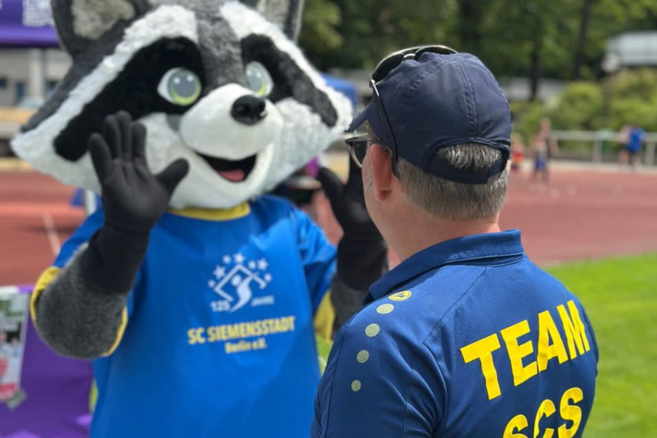 Ein Maskottchen in einem blauen T-Shirt interagiert mit einem Mann im Teamshirt auf einer Sportveranstaltung.