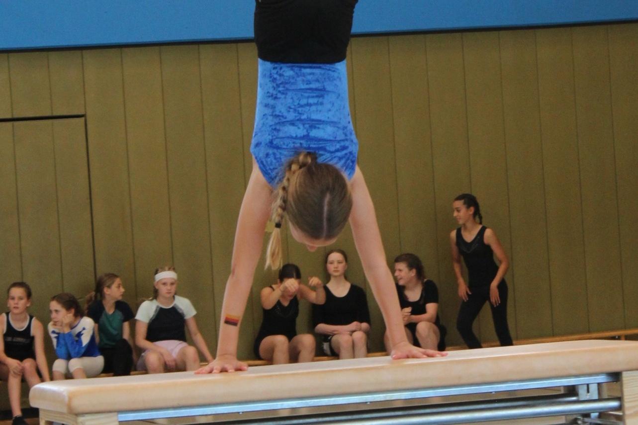 Mädchen übt eine Handstandübung auf einem Barren, während andere Jugendliche zuschauen.
