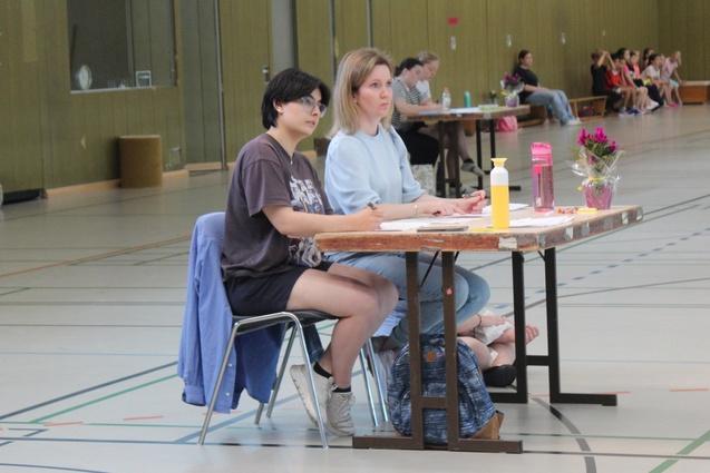Zwei Frauen sitzen an einem Tisch in einer Halle, mit Notizen und Getränken vor sich, im Hintergrund sind weitere Personen zu sehen.