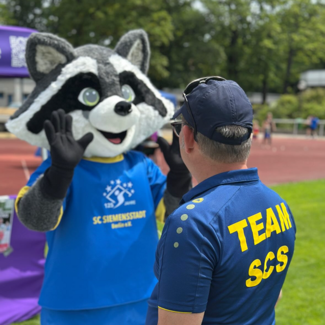 Ein Maskottchen in einem blauen T-Shirt interagiert mit einem Mann im Teamshirt auf einer Sportveranstaltung.
