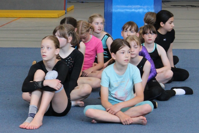 Gruppe von Mädchen in Sportkleidung, sitzend auf dem Boden in einer Turnhalle, aufmerksam und bereit für eine Übung.