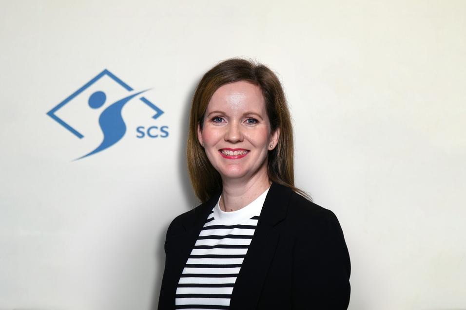 Portr&auml;t einer Frau mit langen Haaren, die in einem schwarzen Blazer und gestreiften Shirt vor einem SCS-Logo steht.