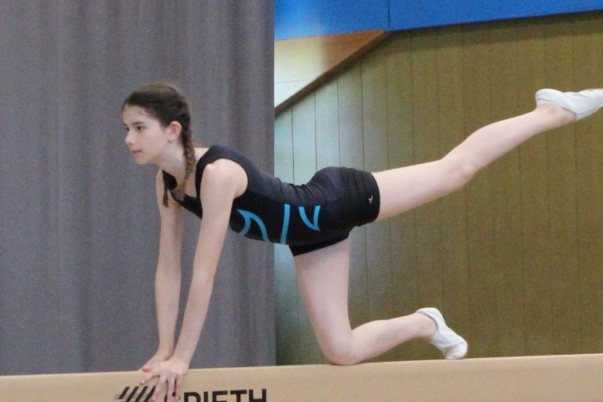 M&auml;dchen in Sportanzug balanciert auf einem Barren, ein Bein nach hinten gestreckt, konzentrierter Gesichtsausdruck.