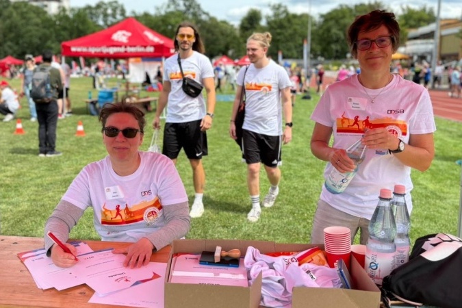 Zwei Personen an einem Tisch mit Materialien und Wasserflaschen, w&auml;hrend im Hintergrund Teilnehmer eines Sportevents zu sehen sind.
