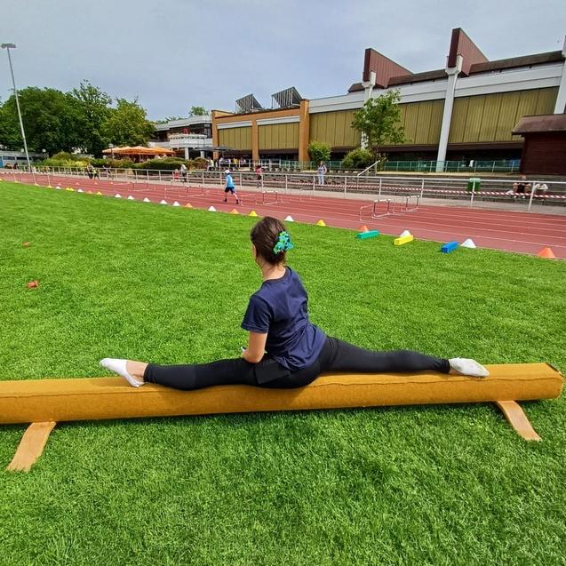 Gymnastin sitzt im Splits auf einem Schwebebalken, umgeben von grünem Rasen und einer Laufbahn im Hintergrund.