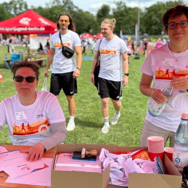Frau mit Sonnenbrille schreibt an einem Tisch, w&auml;hrend zwei M&auml;nner im Hintergrund vorbeigehen, alle in Sportkleidung.