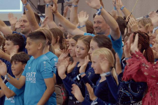 Gruppe von Kindern und Erwachsenen in blauen T-Shirts, die begeistert applaudieren und lächeln.