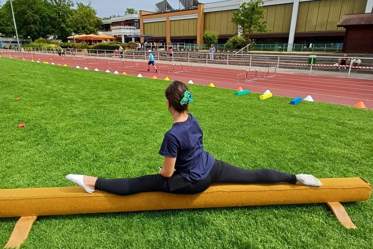 Gymnastin sitzt im Splits auf einem Schwebebalken, umgeben von grünem Rasen und einer Laufbahn im Hintergrund.