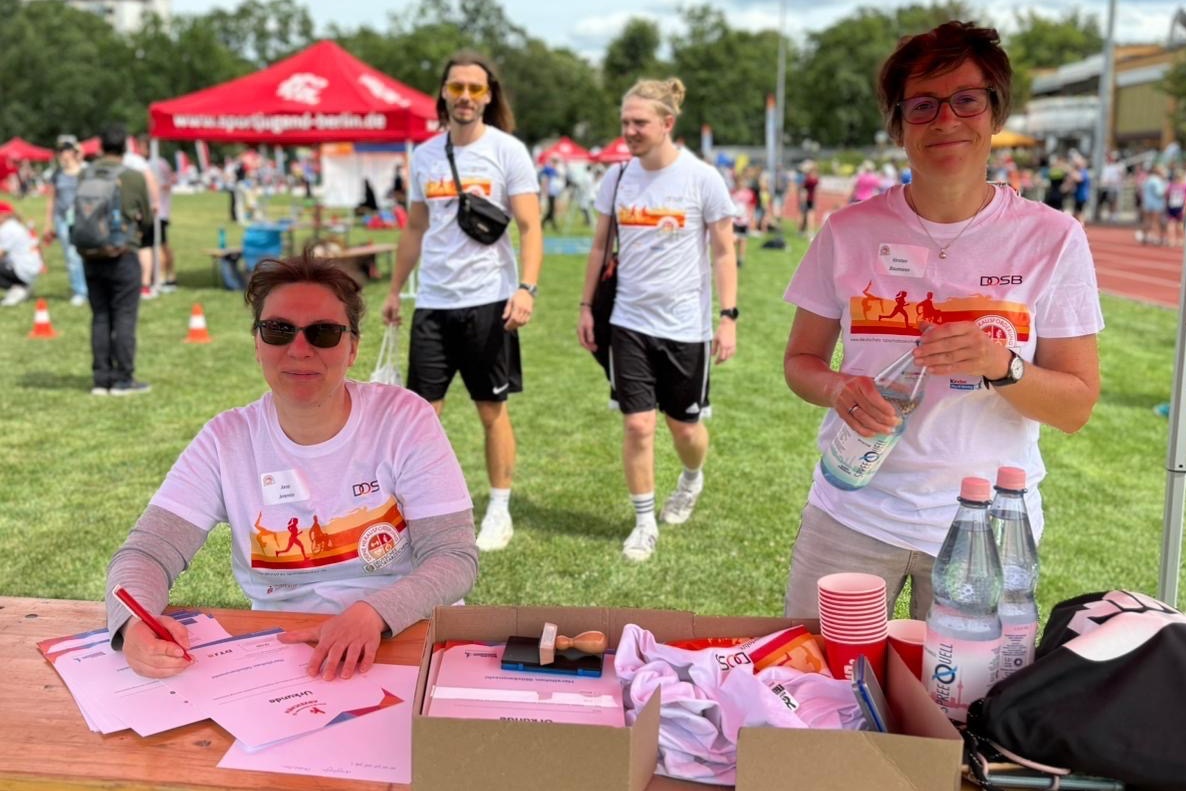 Frau mit Sonnenbrille schreibt an einem Tisch, w&auml;hrend zwei M&auml;nner im Hintergrund vorbeigehen, alle in Sportkleidung.