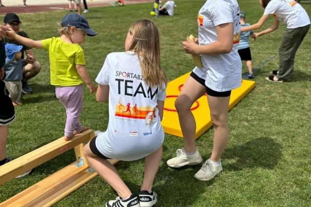 Kinder und Betreuer auf einer Wiese, ein Mädchen steht an einem Balancierbalken, mit Team-Shirts des Sportabzeichens.