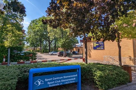 Blauer Hinweisboard des Sport Centrums Siemensstadt mit Parkplatz und B&auml;umen unter klarem Himmel im Hintergrund.