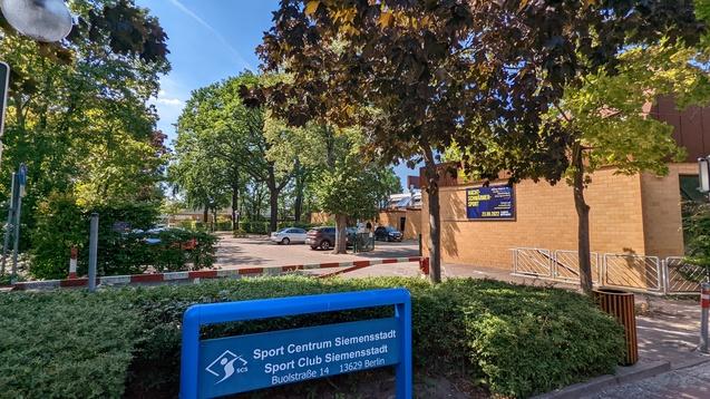 Blauer Hinweisboard des Sport Centrums Siemensstadt mit Parkplatz und Bäumen unter klarem Himmel im Hintergrund.