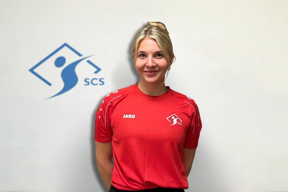 Porträt einer lächelnden Frau in rotem Sportshirt mit Logo, vor einem hellen Hintergrund mit dem SCS-Logo.