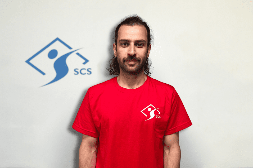 Mann mit lockigem Haar trägt ein rotes T-Shirt mit dem Logo von SCS vor einer hellen Wand.