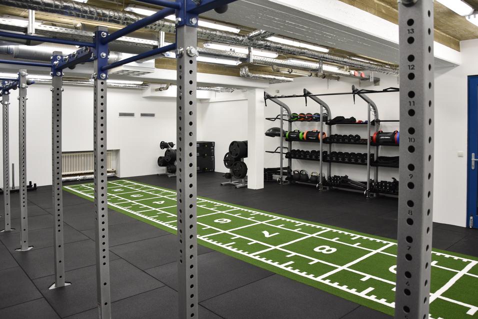 Fitnessraum mit Trainingsger&auml;ten, einer gr&uuml;nen &Uuml;bungsfl&auml;che und Gewichten an Regalen.