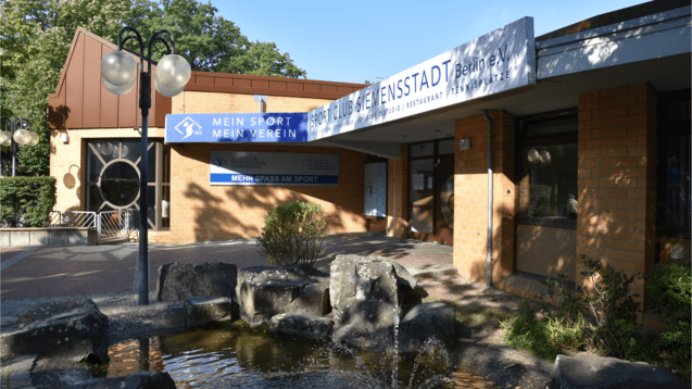 Eingang des Sportclubs Siemenstadt in Berlin mit einem Wasserspiel und beschilderten Gebäudefronten.