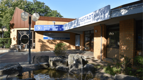 Eingang des Sportclubs Siemenstadt in Berlin mit einem Wasserspiel und beschilderten Gebäudefronten.