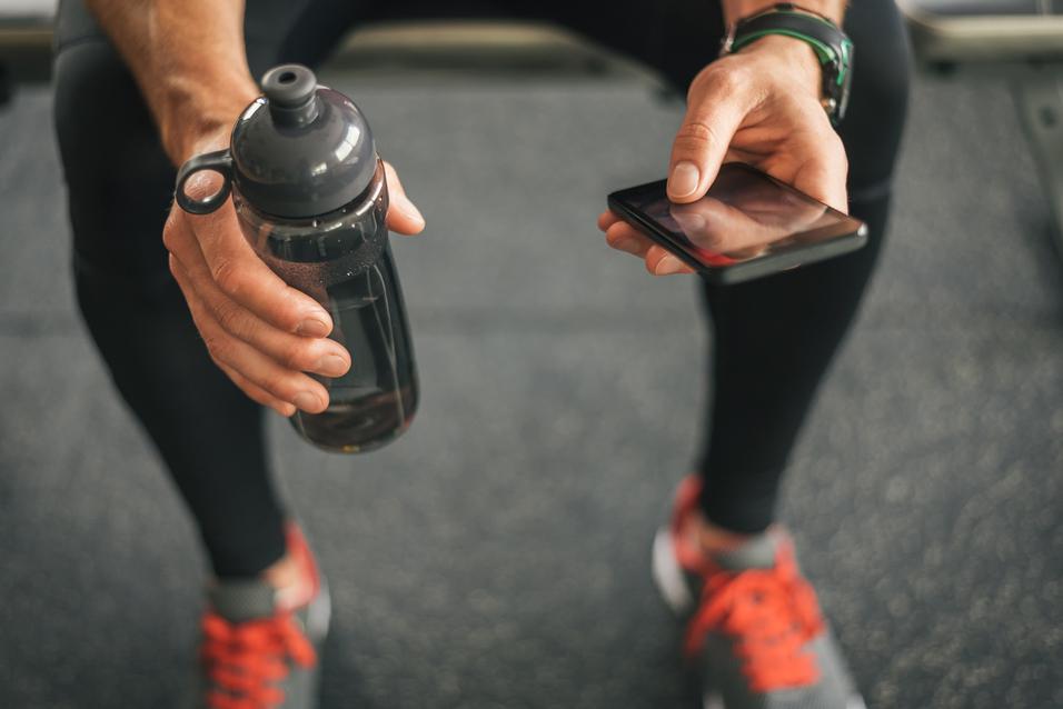 Mann h&auml;lt eine Wasserflasche in einer Hand und ein Smartphone in der anderen, sitzt auf einem Fitnessger&auml;.