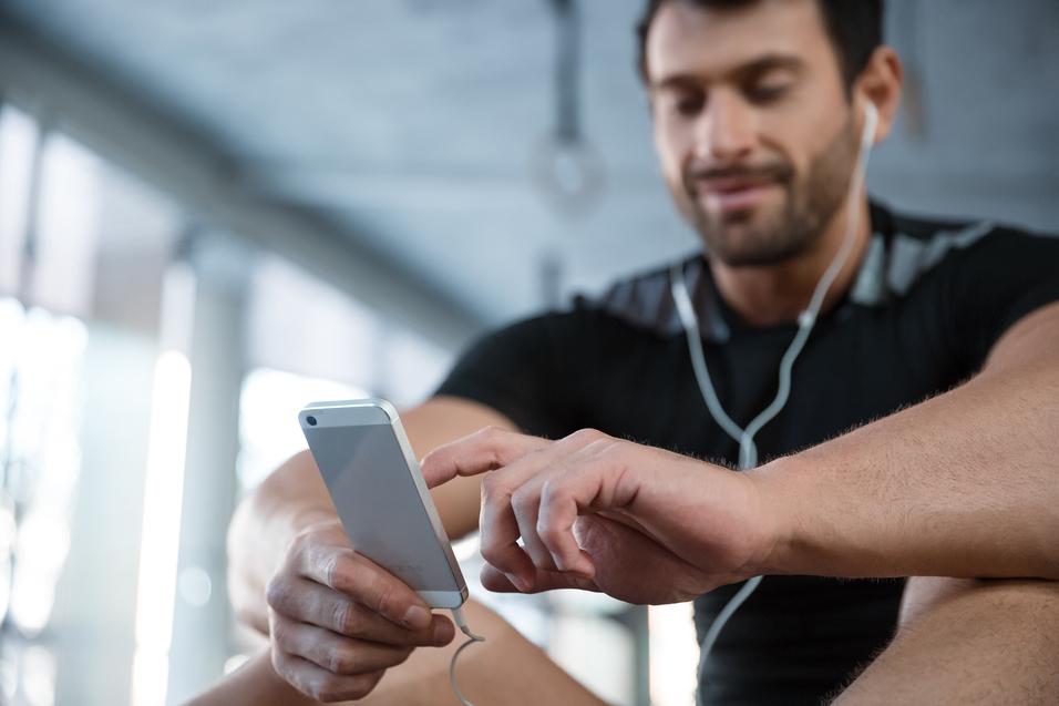 L&auml;chelnder Mann in Sportkleidung sitzt und tippt auf sein Smartphone, w&auml;hrend er Kopfh&ouml;rer tr&auml;gt.