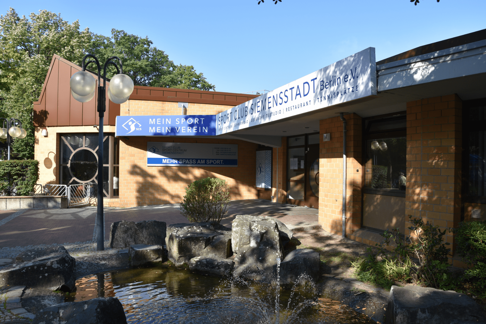 Eingang des Sportclubs Siemenstadt in Berlin mit einem Wasserspiel und beschilderten Geb&auml;udefronten.