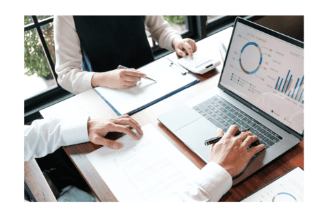 Zwei H&auml;nde &uuml;ber Tabellen und Diagrammen auf einem Tisch, mit Laptop und Taschenrechner im Hintergrund.