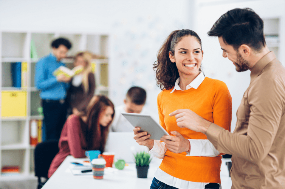 Zwei Menschen stehen in einem modernen Büro und schauen lächelnd auf ein Tablet, während im Hintergrund andere arbeiten.