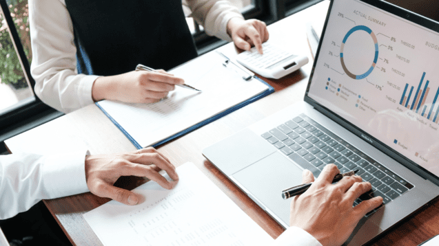 Zwei Hände über Tabellen und Diagrammen auf einem Tisch, mit Laptop und Taschenrechner im Hintergrund.