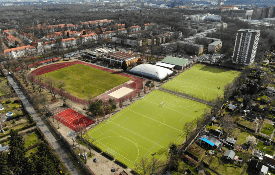 Luftaufnahme eines Sportgeländes mit verschiedenen Sportfeldern, darunter eine Laufbahn, Fußball- und Tennisplätze.