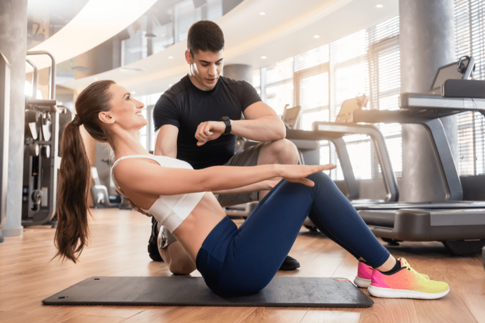 Fitness-Trainer unterstützt lächelnde Frau bei Bauchmuskelübung auf einer Matte im modernen Fitnessstudio.
