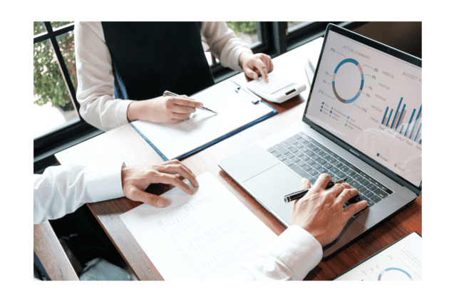 Zwei H&auml;nde &uuml;ber Tabellen und Diagrammen auf einem Tisch, mit Laptop und Taschenrechner im Hintergrund.