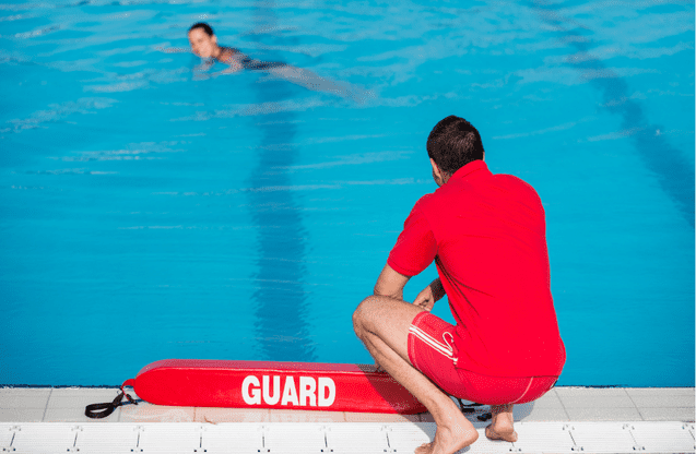 Rettungsschwimmer in roter Uniform beobachtet eine schwimmende Person im Becken. Auf dem Rand liegt ein Rettungsring.