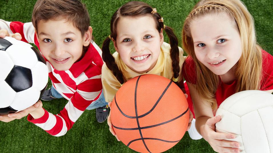 Drei Kinder halten Bälle: einen Fußball, einen Basketball und einen Volleyball, vor einem grünen Rasenhintergrund.