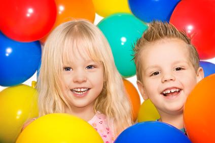 Zwei l&auml;chelnde Kinder stehen zwischen bunten Luftballons in Blau, Rot, Gelb und Gr&uuml;n.