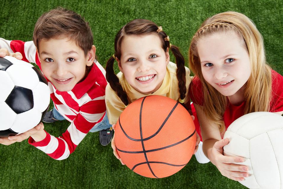 Drei Kinder halten B&auml;lle: einen Fu&szlig;ball, einen Basketball und einen Volleyball, vor einem gr&uuml;nen Rasenhintergrund.