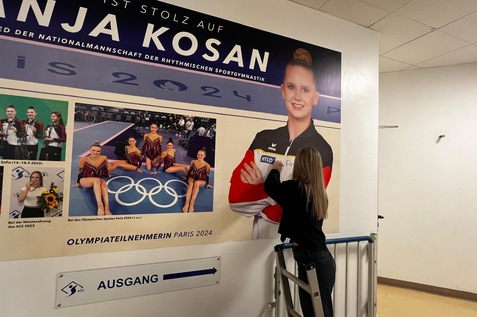 Frau auf einer Leiter, die ein Bild von Anja Kosan, einer Sportlerin, an einer Wand anbringt. Olympische Ringe sichtbar.