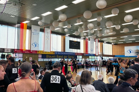 Tanzturnier in einer Halle mit Teilnehmern und Zuschauern, dekoriert mit wei&szlig;en Lampions und bunten Bannern.