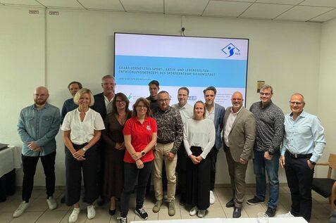 Gruppenfoto von 14 Personen bei einer Pr&auml;sentation zu einem Sportentwicklungsprojekt in einem modernen Raum.