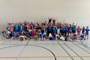 Gruppenfoto von &uuml;ber 70 Kindern in blauen T-Shirts, die freudig in einer Sporthalle posieren.