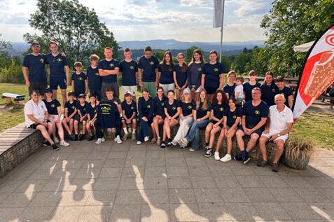 Gruppenfoto von 24 Personen in blauen T-Shirts, im Freien mit B&auml;umen und Blick auf die Landschaft.
